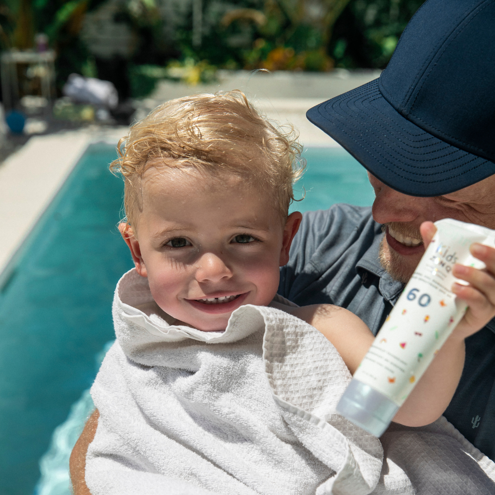 A smiling child wrapped in a towel is held by an adult near a pool, while holding a bottle of Kids Zinq™ SPF 60 sunscreen